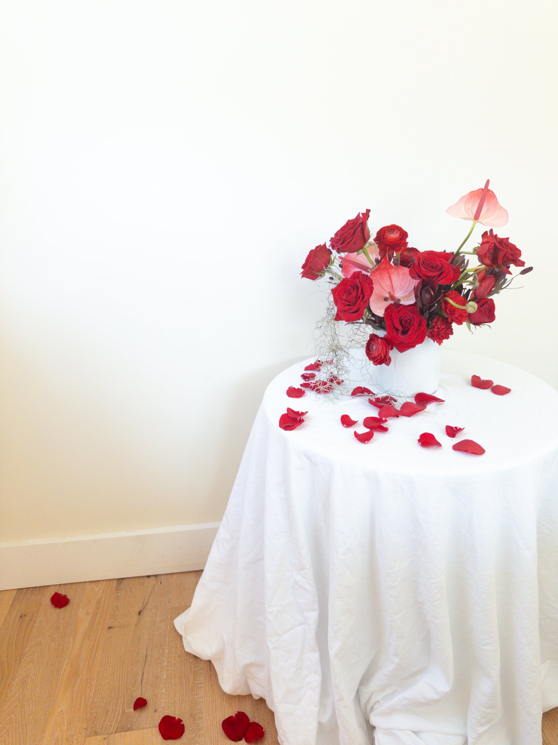 Floral arrangement on a table with red rose petals scattered on a white sheet and floor.
